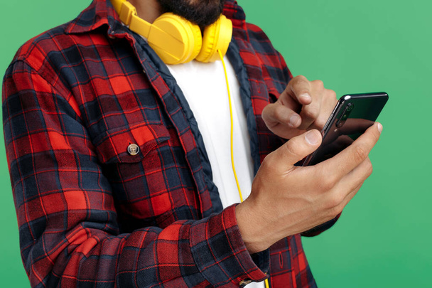 Close-up image of bearded hipster young man in plaid shirt wearing headphones use smartphone over green background - Фото, изображение