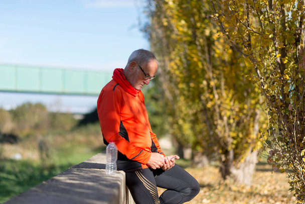 Vista lateral de un hombre corredor de alto nivel apoyado en la cerca mientras prueba el ejercicio en un teléfono móvil al aire libre en un día soleado
 - Foto, imagen