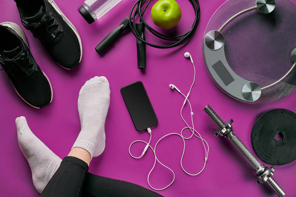 Flat lay gym equipment such as jump rope, bottle of waters, smartphone with headphone, apple and human legs in socks on color background.. Fitness concept - Valokuva, kuva