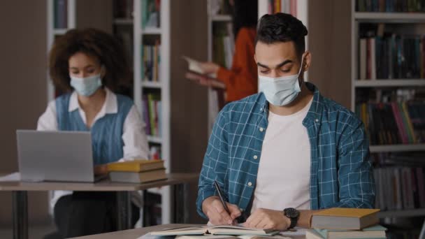 Μαθητές στη βιβλιοθήκη νεαρός Ινδός με προστατευτική μάσκα κάθεται στο γραφείο κάνοντας την προετοιμασία για την εξέταση σημειώσεις γραφής χαρούμενος άνθρωπος κοιτάζοντας κάμερα δείχνει τον αντίχειρα μέχρι χειρονομία έγκρισης υπογράψει εξαιρετικό σήμα - Πλάνα, βίντεο