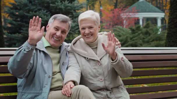 Caucasian married couple family retired 60s people man woman waving hands hello goodbye gesture elderly grandparents sit on bench in autumn park lovely talking gesticulate having pleasant conversation - Materiaali, video
