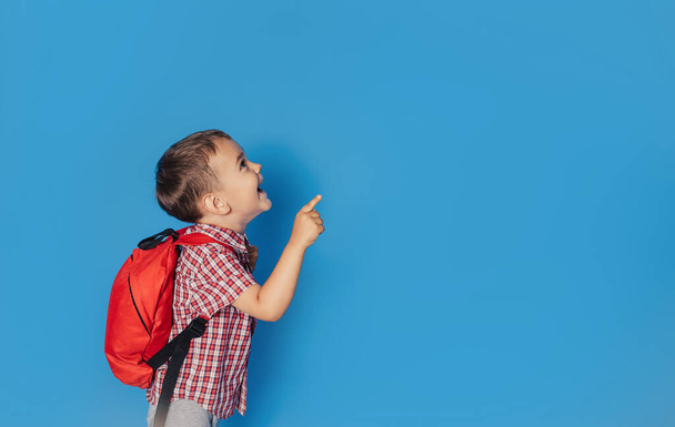 Veselý usmívající se malý brunetka chlapec s batohem se baví na pozadí modré zdi a ukazuje na váš text. Koncept školy. zpět do školy. - Fotografie, Obrázek