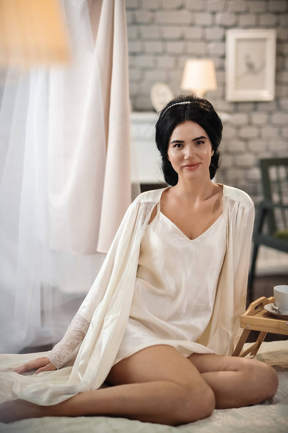 hermosa mujer joven en vestido blanco sentado en la cama en el dormitorio - Foto, imagen
