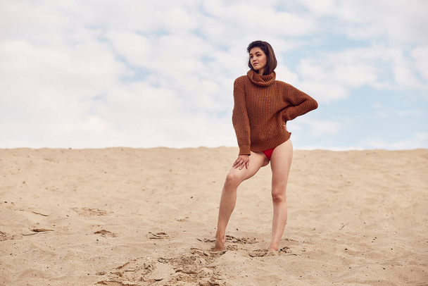 Beautiful woman with windy hair standing on sandy beach. Stylish young sexy female in knitted sweater and bare legs relaxing on coast. - 写真・画像
