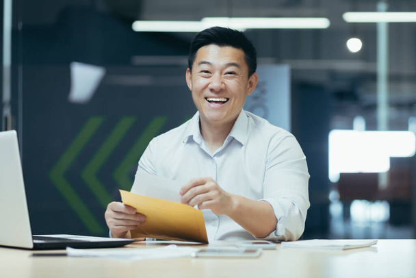 Portrait of successful Asian investor, man puts money money cash in envelope, smiling and looking at camera, working in modern office - Foto, Imagen