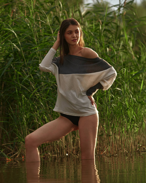 Attractive young brunette in a sweater and and black pantians posing on the reeds beach. Beach fashion, seductive woman resting near the water - Φωτογραφία, εικόνα