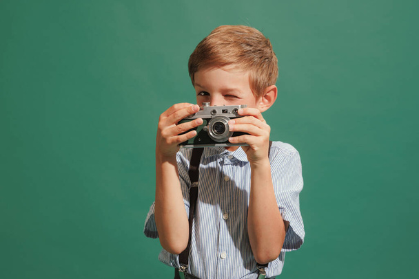 Taking picture with retro phote camera. One preschool boy, kid wearing retro style outfit isolated on dark green background. Concept of child emotions, facial expression, fashion and ad. - Foto, immagini