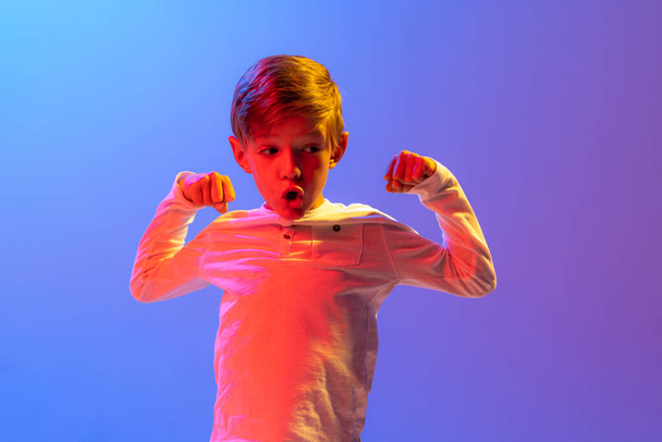 Strength. Little athlete. Studio shot of stylish little boy, kid posing isolated on gradient blue-purple background in neon light. Concept of emotions, facial expression, fashion, hobbies, active - Fotó, kép