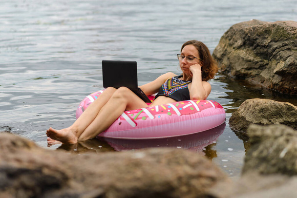 A workaholic woman works in nature. A freelancer works on a laptop while floating on a lake on a rubber ring. - Foto, Imagem