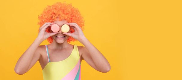 happy child in swimsuit wearing orange curly wig hair holding french macaron on yellow background, sweet tooth. - Photo, Image