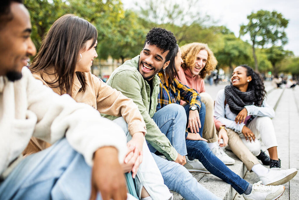 Happy group of trendy young people laughing sitting in city street. Millennial diverse student friends having fun together outdoor - Foto, Imagen
