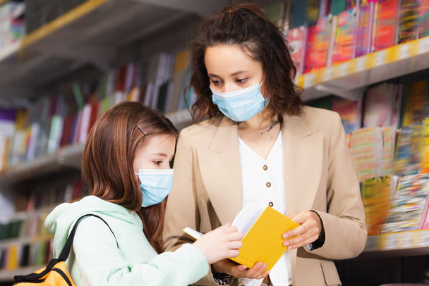 woman in medical mask showing new notebook to daughter in stationery store - 写真・画像