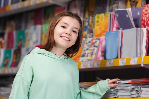happy child looking at camera near blurred rack with colorful notebooks - Фото, изображение