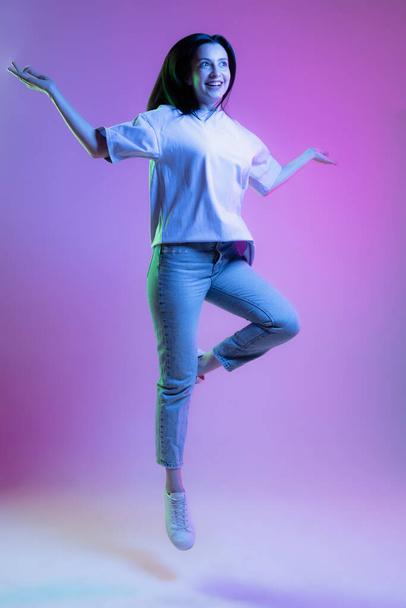 Jumping, flying. Portrait of adorable girl, young woman with long glossy dark hair isolated on purple background in neon light. Concept of beauty, art, fashion, youth, sales and ads. Happiness - 写真・画像