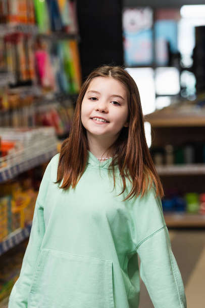 happy schoolgirl smiling at camera in blurred stationery shop - Foto, imagen