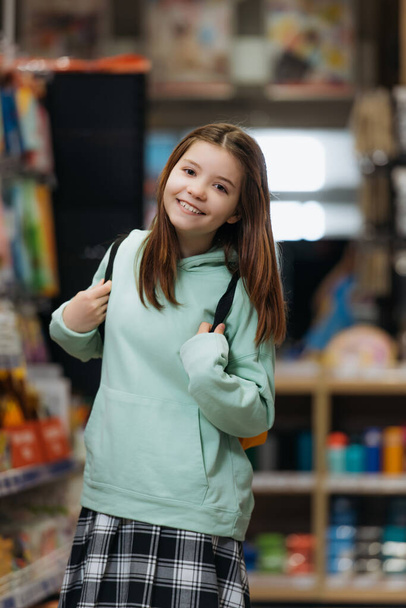 pleased schoolgirl with backpack smiling at camera in stationery shop - Foto, Imagem