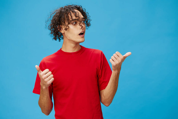 Excited shocked funny curly tanned Caucasian guy in basic red t-shirt show thumb up gesture Like posing isolated on blue background. Lifestyle and Emotions concept. Good offer with copy space for ad - Photo, Image