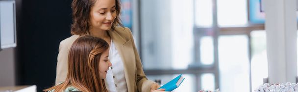smiling woman showing new copybook to daughter in stationery store, banner - Foto, Imagem