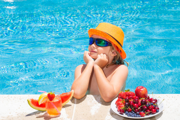 A kisgyerek a medencénél gyümölcsöt eszik és limonádé koktélt iszik. Nyári gyerekek nyaralás koncepció - Fotó, kép