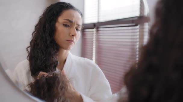 Worried upset young woman standing in bathroom in bathrobe reflected in mirror looking at split ends brittle damaged dry hair unhappy girl feeling frustration about unhealthy long curly fragile hair - Záběry, video