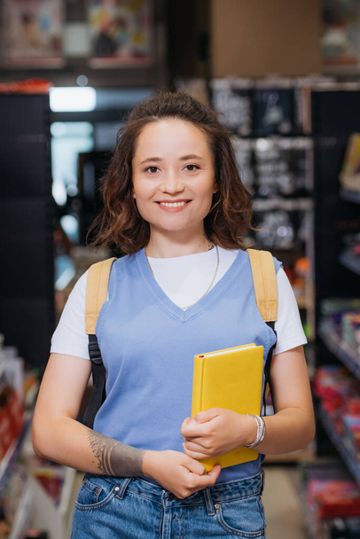 young woman with new copybook smiling at camera in stationery shop - Фото, изображение