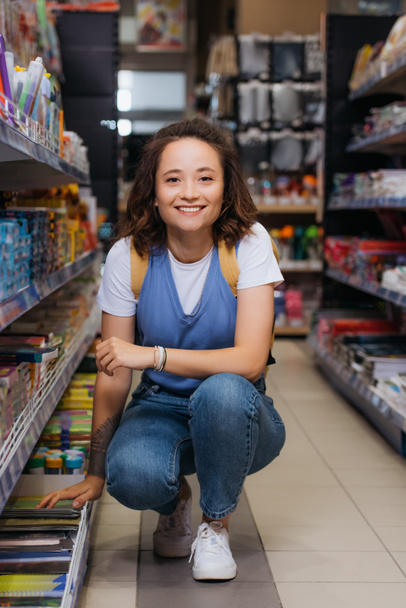 cheerful young woman in jeans looking at camera near racks in stationery shop - Foto, afbeelding