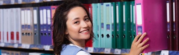 happy woman taking folder from rack in stationery store and looking at camera, banner - 写真・画像