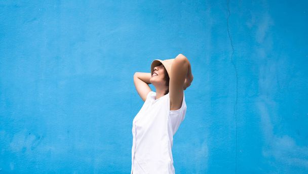 Женщина в белой рубашке, стоящей перед синей стеной, поднимите руку, дотроньтесь до шляпы, повернитесь лицом вверх и улыбнитесь, человеческие эмоции - Фото, изображение