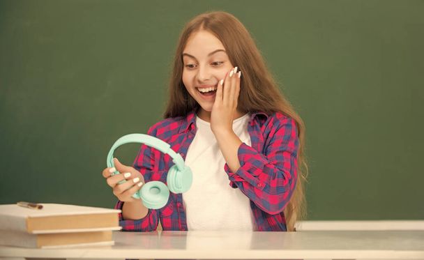 surpresa adolescente menina segurar fones de ouvido modernos no ensino médio no quadro negro, escola. - Foto, Imagem