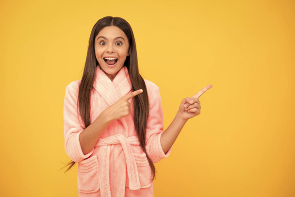 щаслива дівчина-підліток в домашніх умовах махровий халат вказує палець на місце для копіювання, напрямок
. - Фото, зображення