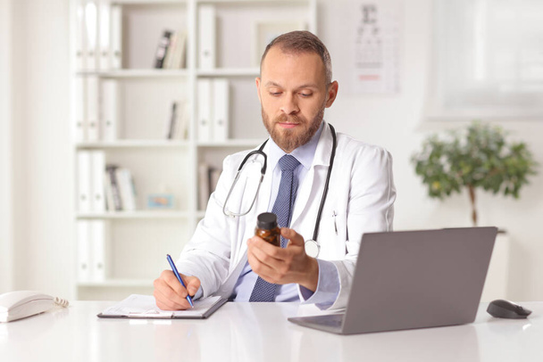 Young male doctor sitting in an office holding a bottle of pills and writing a document - Foto, imagen