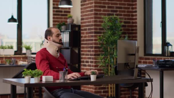 Männlicher Angestellter mit Schaumstoffkragen, der mit dem Computer im Büro am Geschäftswachstum arbeitet. Manager trägt Halsband, um sich nach Knochenbruch und Wirbelverletzung zu erholen. Stativschuss. - Filmmaterial, Video