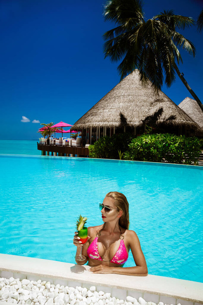 Beautiful female model in pink bikini is standing in the pool and holding colorful fruity drink. Sexy woman in sequin bikini is posing in the pool on Maldives island. Luxury travel. Vacations. - Photo, Image