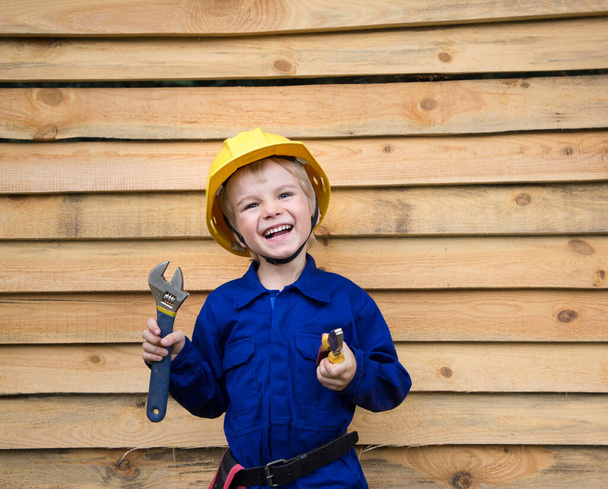 Porträt eines netten wissbegierigen Jungen im Alter von 6 Jahren in blauer Overalls-Uniform und Bauhelm mit Werkzeug in der Hand auf einem hölzernen Hintergrund. Reparaturassistent, kleiner Helfer. Sei wie Papa - Foto, Bild