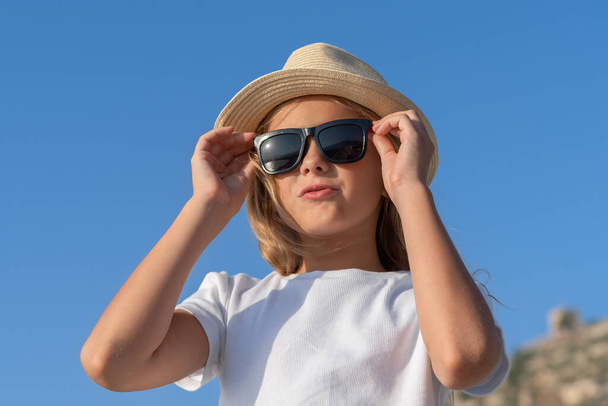 A young girl in a hat and sunglasses adjusts her glasses with two hands against the blue sky. - 写真・画像