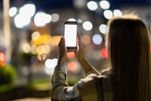 Mladá žena drží mobilní telefon s prázdným bílým displejem na pozadí světlé noční ulice města - Fotografie, Obrázek