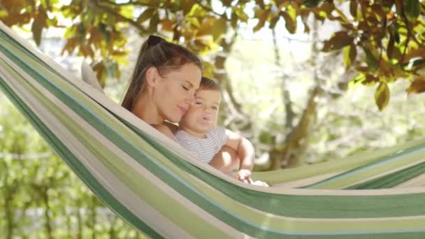 smiling happy mother hugs and cuddle her little baby son, relaxing  lying together in the hammock. Child plays with pacifier, in the home garden on a sunny summer day, joyful family childhood concept - Imágenes, Vídeo