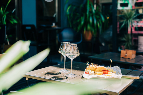 Une table en bois dans un restaurant avec une assiette de fromage, raisin, pain au miel et vin blanc. Verres à vin et fromage servis pour une fête dans un bar flou ou un restaurant sur la terrasse lors d'une journée d'été ensoleillée. - Photo, image