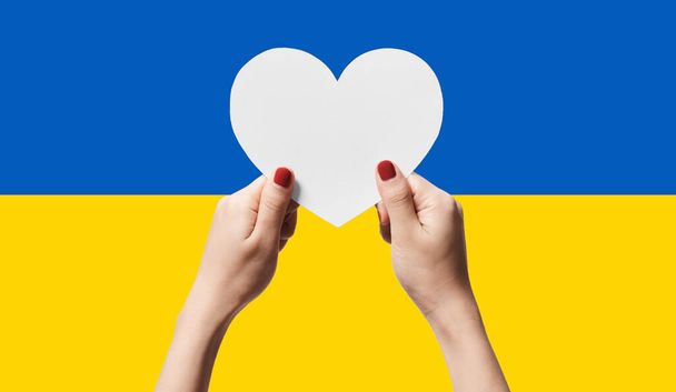 Women hands holding heart shape paper on Ukraine flag color background. Stay with ukraine symbol. Hand heart love gesture with ukrainian flag background. - Photo, Image