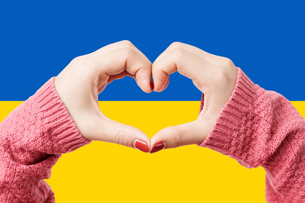 Kobiety ręce tworząc kształt serca na tle flagi Ukrainy kolor. Trzymaj się ukraińskiego symbolu. Ręka serce miłość gest z ukraińskiej flagi tle. - Zdjęcie, obraz