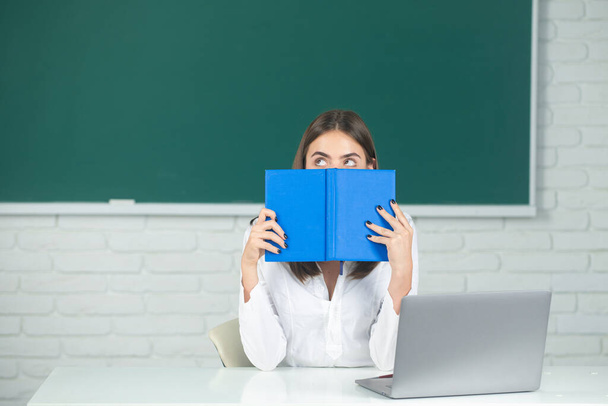 Portrait of a young, confident and attractive female student studying in school classroom. Female college student hiding behind an open book and looking away. Girl covering face with book - Φωτογραφία, εικόνα