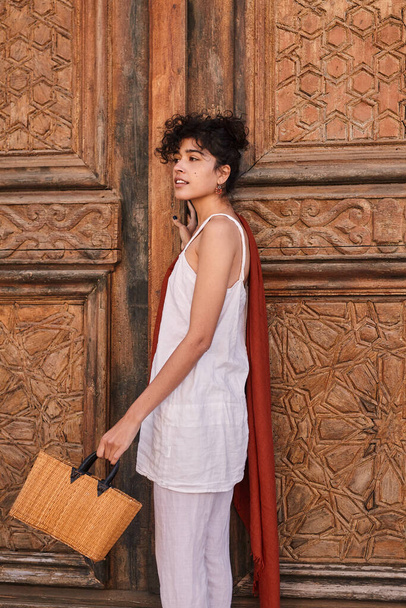 Beautiful brunette woman in fashionable clothes posing near wooden door with bag. Female fashion and summer concept - Φωτογραφία, εικόνα