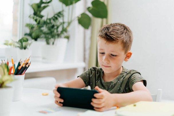 School kid boy studying at home with mobile smart phone and doing homework. Back to school. Thinking caucasian child with cellphone, pencil and training books. Distance learning online education. - Φωτογραφία, εικόνα