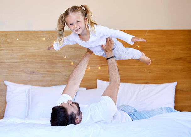 Adorable little girl bonding with her single father at home and pretending she can fly. Caucasian single parent holding and lifting his daughter in the air. Smiling child playing in the bedroom. - Fotoğraf, Görsel