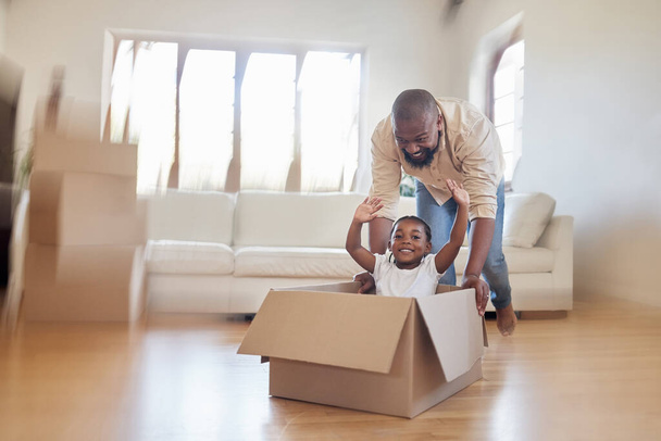 Cheerful african american father pushing excited little girl around in carton box, riding around and having fun in living room. Happy dad and daughter enjoying moving day at their new home. - Фото, изображение
