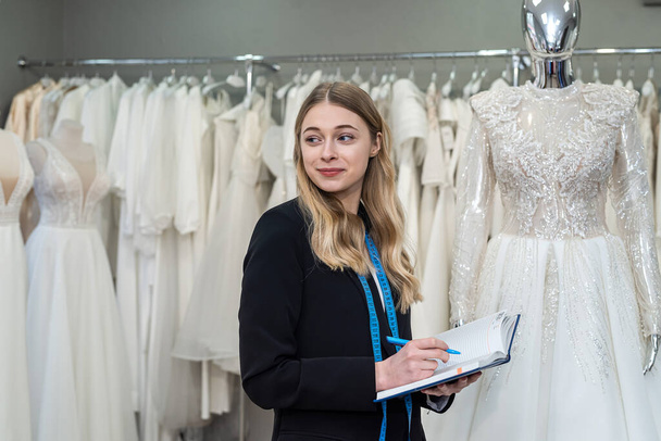 hübsche Designerin oder Verkäuferin macht sich Notizen in der Nähe des Kleides im Geschäft für Hochzeitsmode.  - Foto, Bild