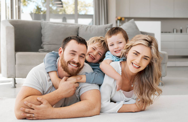 Portrait of smiling caucasian family of four relaxing on floor in lounge at home. Playful sons lying and clinging on carefree loving parents backs while bonding and spending fun quality time together. - Foto, imagen