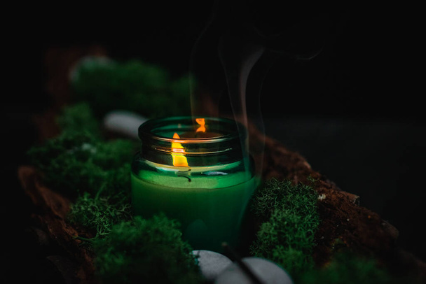 A white lit candle in a glass jar with flowing smoke, green moss and gray stones in the bark of a tree lies diagonally against a black background, close-up side view. Esoteric concept, dark style. - Zdjęcie, obraz