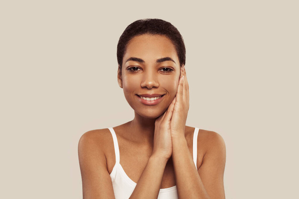 Güzel surat. Doğal makyajı ve sağlıklı deri portresi olan Afrikalı Amerikalı bir kadın. Güzel bir model, beyaz arka planda parlayan hidratlı cilde dokunuyor. Cilt bakımı kavramı - Fotoğraf, Görsel