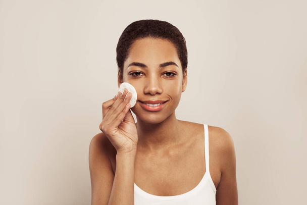 Mladý krásný africký americký žena čištění její tvář z make-upu s bílou bavlněnou podložky na bílém pozadí - Fotografie, Obrázek